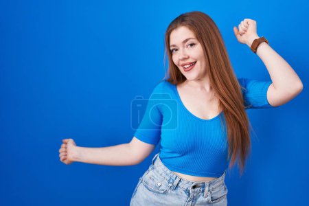 Téléchargez les photos : Femme rousse debout sur fond bleu dansant heureux et joyeux, souriant décontracté et confiant écoutant de la musique - en image libre de droit