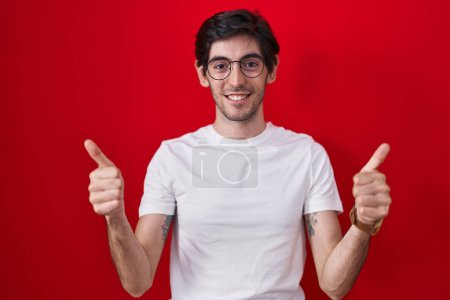 Foto de Joven hombre hispano de pie sobre el signo de éxito de fondo rojo haciendo gesto positivo con la mano, pulgares arriba sonriendo y feliz. expresión alegre y gesto ganador. - Imagen libre de derechos
