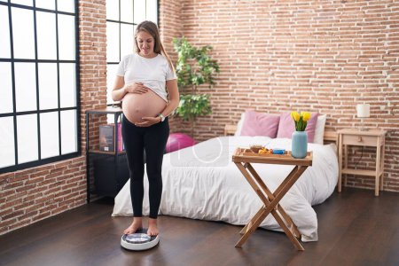 Foto de Mujer embarazada joven de pie en la máquina de pesaje tocando el vientre en el dormitorio - Imagen libre de derechos