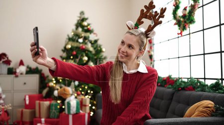Photo for Young blonde woman make selfie by smartphone sitting on sofa by christmas tree at home - Royalty Free Image