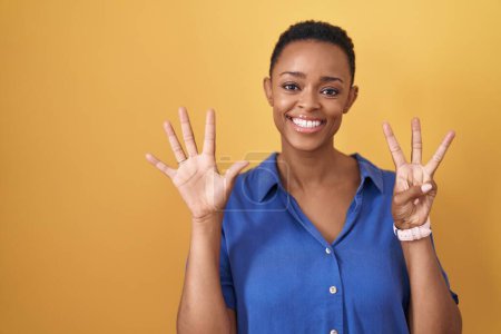 Foto de Mujer afroamericana de pie sobre fondo amarillo mostrando y señalando hacia arriba con los dedos número ocho mientras sonríe confiado y feliz. - Imagen libre de derechos