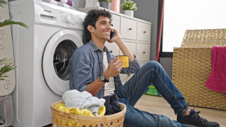 Téléchargez les photos : Jeune homme hispanique parlant sur smartphone boire du café en attendant la machine à laver à la buanderie - en image libre de droit
