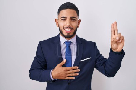 Téléchargez les photos : Jeune homme hispanique portant un costume d'affaires et cravate souriant jurant avec la main sur la poitrine et les doigts vers le haut, faisant un serment de fidélité promesse - en image libre de droit