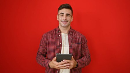 Cheerful young hispanic man radiating confidence, smiling over an isolated red background while casually standing, engrossed in his touchpad.