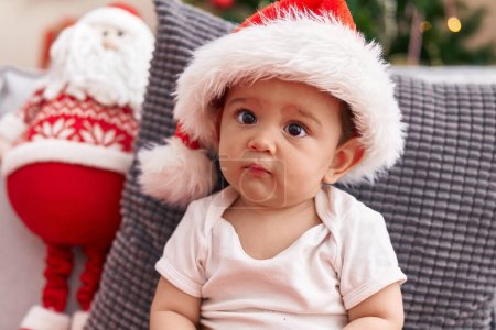 Photo for Adorable hispanic baby wearing christmas hat sitting on sofa at home - Royalty Free Image
