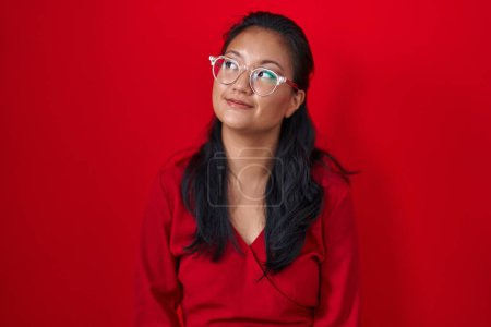 Téléchargez les photos : Asiatique jeune femme debout sur fond rouge souriant regardant vers le côté et regardant loin en pensant. - en image libre de droit