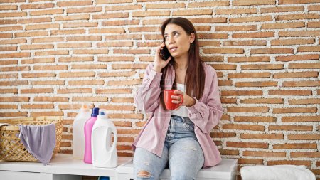 Téléchargez les photos : Jeune belle femme hispanique parlant sur smartphone boire du café en attendant la machine à laver à la buanderie - en image libre de droit