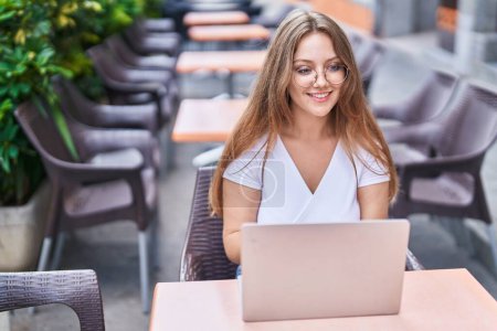 Téléchargez les photos : Jeune femme blonde utilisant un ordinateur portable assis sur la table à la terrasse de café - en image libre de droit
