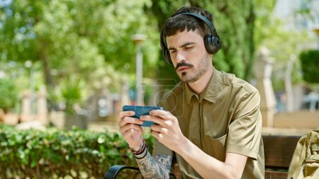Téléchargez les photos : Jeune homme hispanique jouant à un jeu vidéo assis sur un banc au parc - en image libre de droit
