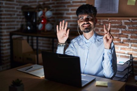 Téléchargez les photos : Jeune homme hispanique avec la barbe travaillant au bureau la nuit montrant et pointant vers le haut avec les doigts numéro sept tout en souriant confiant et heureux. - en image libre de droit