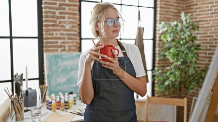Foto de Joven mujer rubia artista mirando dibujo celebración taza de café en el estudio de arte - Imagen libre de derechos