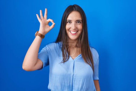 Téléchargez les photos : Young brunette woman standing over blue background smiling positive doing ok sign with hand and fingers. successful expression. - en image libre de droit