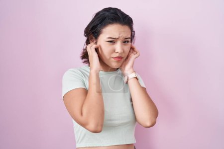 Foto de Mujer joven hispana de pie sobre fondo rosa cubriendo las orejas con los dedos con expresión molesta por el ruido de la música alta. concepto de sordo. - Imagen libre de derechos