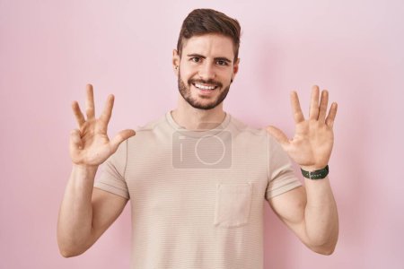 Foto de Hombre hispano con barba de pie sobre fondo rosa mostrando y señalando hacia arriba con los dedos número nueve mientras sonríe confiado y feliz. - Imagen libre de derechos
