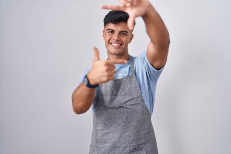 Téléchargez les photos : Jeune homme hispanique portant tablier sur fond blanc souriant faisant cadre avec les mains et les doigts avec le visage heureux. concept de créativité et photographie. - en image libre de droit