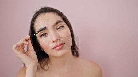 Foto de Joven hermosa mujer hispana aplicando maquillaje en las pestañas sobre fondo rosa aislado - Imagen libre de derechos