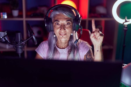 Téléchargez les photos : Femme d'âge moyen avec des tatouages jouant à des jeux vidéo portant un casque souriant avec une idée ou une question pointant du doigt avec un visage heureux, numéro un - en image libre de droit