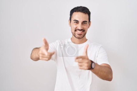Foto de Hombre hispano guapo de pie sobre fondo blanco señalando los dedos a la cámara con la cara feliz y divertida. buena energía y vibraciones. - Imagen libre de derechos