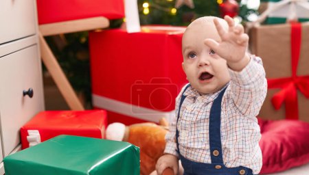 Photo for Adorable caucasian baby sitting on floor by christmas tree with hand raised up at home - Royalty Free Image