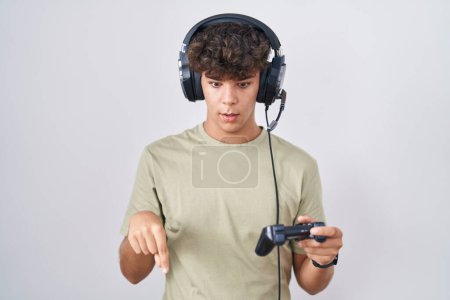 Photo for Hispanic teenager playing video game holding controller pointing down with fingers showing advertisement, surprised face and open mouth - Royalty Free Image