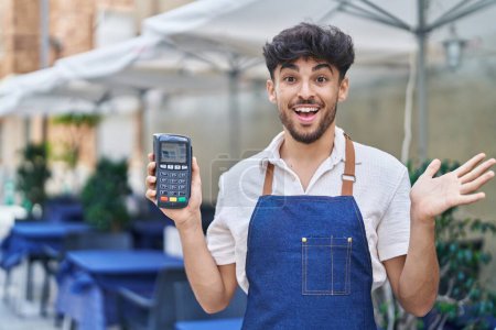 Photo for Arab man with beard wearing waiter apron at restaurant terrace holding dataphone celebrating achievement with happy smile and winner expression with raised hand - Royalty Free Image