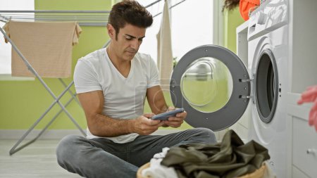 Téléchargez les photos : Jeune homme hispanique jouant jeu vidéo laver les vêtements à la buanderie - en image libre de droit