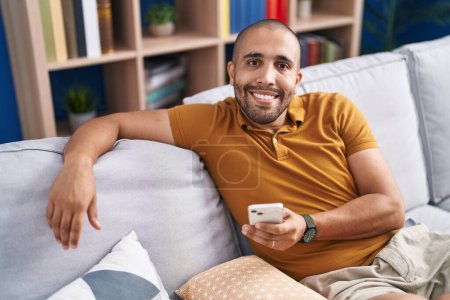 Téléchargez les photos : Jeune homme latino utilisant smartphone assis sur le canapé à la maison - en image libre de droit