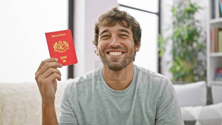 Foto de Joven guapo sonriendo de alegría, sentado en un sofá en casa, sosteniendo su pasaporte malayo, un epítome de la confianza y la felicidad casuales. - Imagen libre de derechos