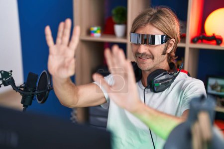 Téléchargez les photos : Jeune homme blond streamer jouer à un jeu vidéo en utilisant des lunettes de réalité virtuelle à la salle de jeu - en image libre de droit