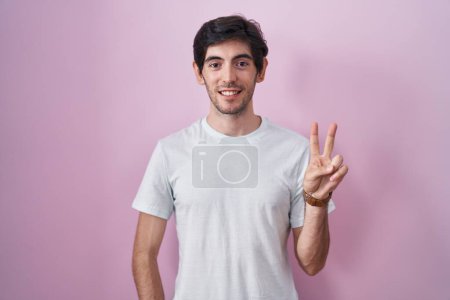 Foto de Young hispanic man standing over pink background showing and pointing up with fingers number two while smiling confident and happy. - Imagen libre de derechos