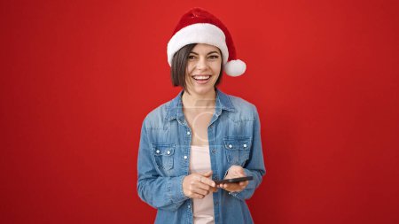 Téléchargez les photos : Jeune femme caucasienne portant un chapeau de Noël en utilisant un smartphone sur fond rouge isolé - en image libre de droit