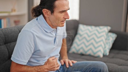 Téléchargez les photos : Homme d'âge moyen souffrant de maux d'estomac assis sur le canapé à la maison - en image libre de droit