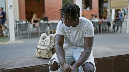 Foto de Hombre afroamericano mirando hacia un lado con expresión seria sentado en el banco en la terraza de la cafetería - Imagen libre de derechos