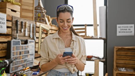 Photo for Smiling young hispanic woman, a beautiful carpenter, confidently texting on her smartphone amid the timber at an indoor carpentry workshop. - Royalty Free Image