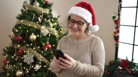 Téléchargez les photos : Femme d'âge moyen avec les cheveux gris en utilisant smartphone par arbre de Noël à la maison - en image libre de droit