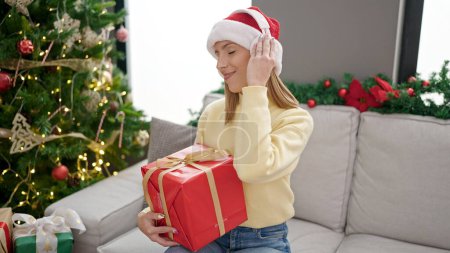Foto de Joven mujer rubia escuchando música sosteniendo regalo por árbol de Navidad en casa - Imagen libre de derechos