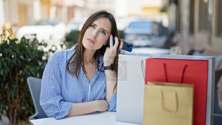 Téléchargez les photos : Jeune belle femme hispanique écoutant un message vocal par smartphone assis sur la table avec des sacs à provisions sur la terrasse du café - en image libre de droit