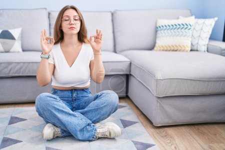 Téléchargez les photos : Jeune femme caucasienne assise par terre dans le salon, détendue et souriante, les yeux fermés, faisant un geste de méditation avec les doigts. concept de yoga. - en image libre de droit