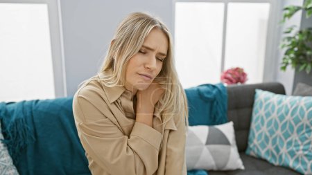 Téléchargez les photos : Inquiète et malheureuse, la jeune femme blonde souffre de douleurs cervicales sévères assise seule sur le canapé à la maison - en image libre de droit