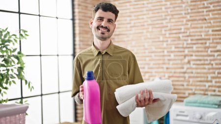 Foto de Joven hombre hispano sonriendo confiado sosteniendo toallas dobladas y botella de detergente en la lavandería - Imagen libre de derechos