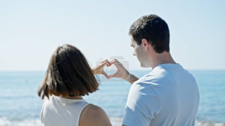 Téléchargez les photos : Beau couple faisant geste de cœur avec les mains debout vers l'arrière au bord de la mer - en image libre de droit