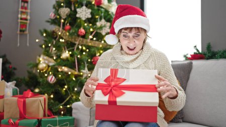 Photo for Mature hispanic woman wearing christmas hat unpacking gift with surprised face at home - Royalty Free Image