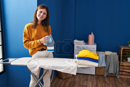 Foto de Joven mujer rubia sonriendo confiada máquina de planchar de limpieza en la sala de lavandería - Imagen libre de derechos