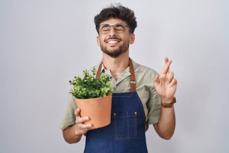 Téléchargez les photos : Homme arabe avec barbe tenant pot de plante verte geste doigt croisé souriant avec espoir et les yeux fermés. chance et concept superstitieux. - en image libre de droit