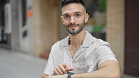 Téléchargez les photos : Jeune homme hispanique pointant vers regarder assis sur le banc souriant à la rue - en image libre de droit