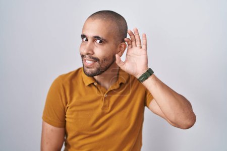 Téléchargez les photos : Homme hispanique avec la barbe debout sur fond blanc souriant avec la main sur l'oreille en écoutant une rumeur ou des commérages. concept de surdité. - en image libre de droit