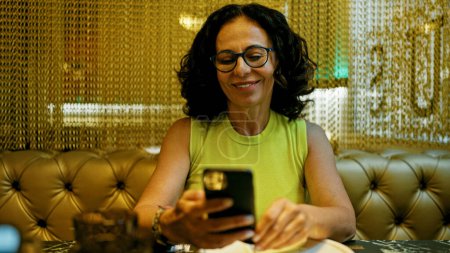 Téléchargez les photos : Femme hispanique du Moyen Âge utilisant un smartphone dans un restaurant - en image libre de droit