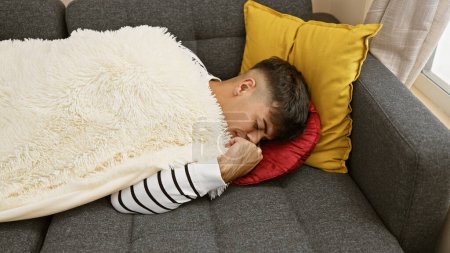 Foto de Hombre hispano joven agotado y guapo encuentra consuelo en una acogedora siesta, acostado en casa en el sofá en un merecido descanso, capturando perfectamente el retrato de relajación interior de un adulto en la sala de estar. - Imagen libre de derechos