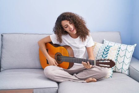 Téléchargez les photos : Jeune belle femme hispanique jouant de la guitare classique assise sur un canapé à la maison - en image libre de droit