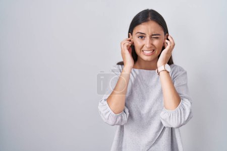 Téléchargez les photos : Jeune femme hispanique debout sur fond blanc couvrant les oreilles avec des doigts avec une expression agacée pour le bruit de la musique forte. concept sourd. - en image libre de droit